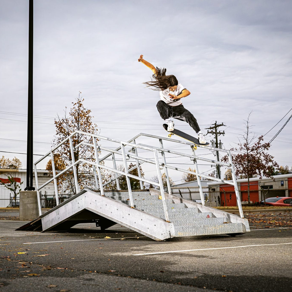 A skateboarder mid-air performs a trick over a metal stair rail in an outdoor urban setting under an overcast sky.