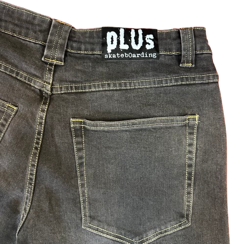 Close-up of the back waistband and pocket area of black denim jeans, showing a black label with white text that reads 'PLUs skateboarding'.