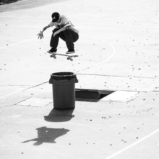 A black and white action photograph of a skateboarder mid-jump over a trash can on an outdoor concrete court.