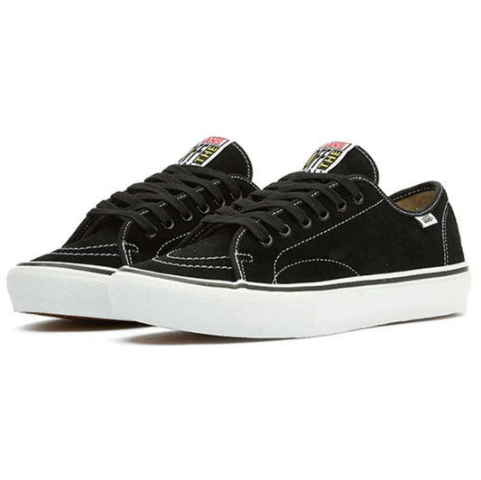 A pair of black suede low-top sneakers with white rubber soles, black laces, and a colorful logo patch on the tongue