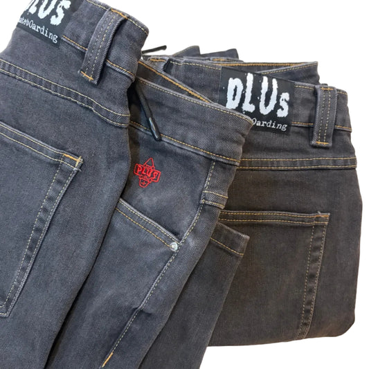 Two pairs of folded dark gray denim jeans laid flat on a light wood floor. One pair has white 'PLUS' branding and the other has a red 'PLUS' logo patch.