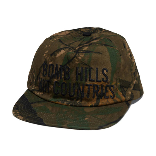 Camouflage cap with 'BOMB HILLS NOT COUNTRIES' text on a white background