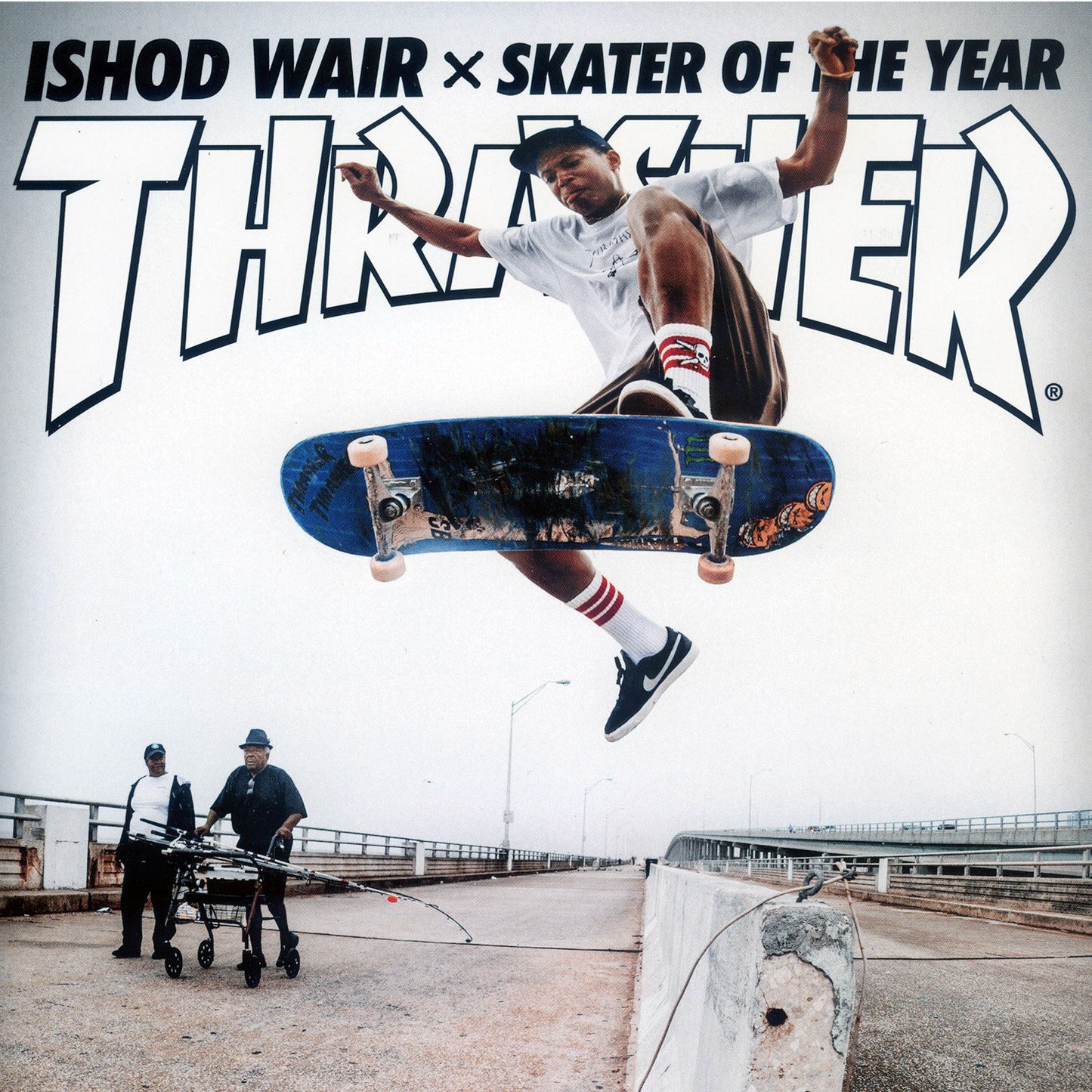 A skateboarder in midair performing a trick over a bridge, with large 'Thriver' text and the words 'Ishod Wair x Skater of the Year' across the top. Two men push a cart in the background on the bridge.