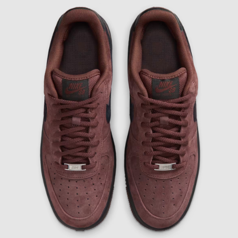 A pair of Nike Air Force 1 Low sneakers in a dark brown suede featuring dark brown laces, a dark brown tongue label with 'Nike SB' branding, and a silver metal lace guard, shown from a top-down view 