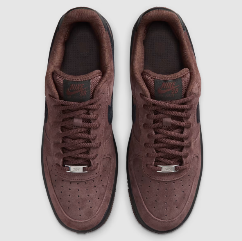 A pair of Nike Air Force 1 Low sneakers in a dark brown suede featuring dark brown laces, a dark brown tongue label with 'Nike SB' branding, and a silver metal lace guard, shown from a top-down view 