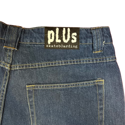 Close-up of the back waistband of dark blue denim jeans, showing a black brand patch with 'pLUs skateboarding' in white text, alongside the back pocket and yellow contrast stitching.