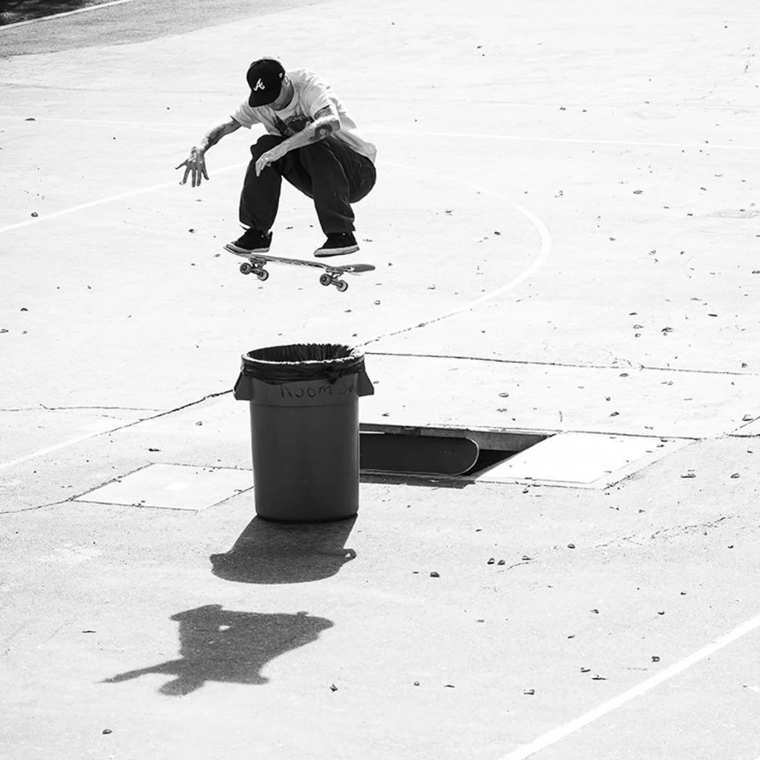 A black and white action photograph of a skateboarder mid-jump over a trash can on an outdoor concrete court.