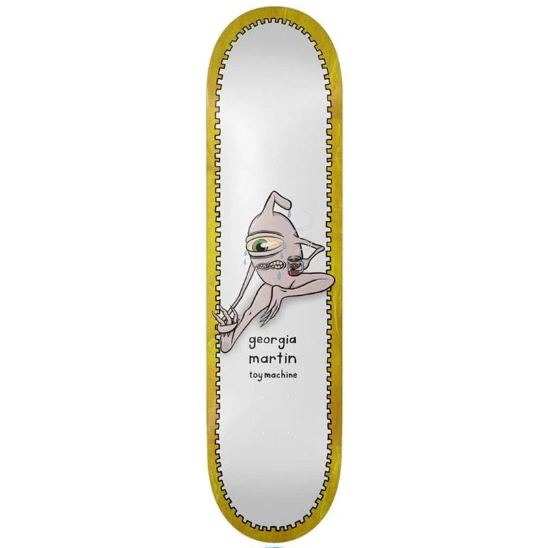 A skateboard deck shown vertically against a plain white background. The deck has a white center with a cartoon alien graphic and the text 'georgia martin toy machines', surrounded by a gold-colored inner border and a black outer edge.