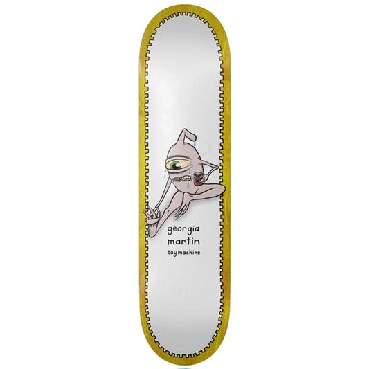 A skateboard deck shown vertically against a plain white background. The deck has a white center with a cartoon alien graphic and the text 'georgia martin toy machines', surrounded by a gold-colored inner border and a black outer edge.