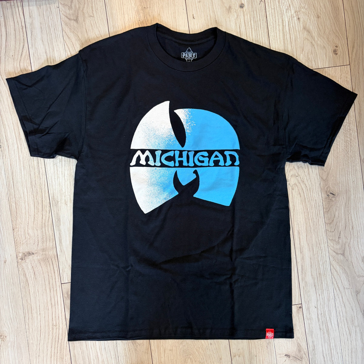 A black short-sleeve t-shirt laid flat on a light wood floor, featuring a large blue and white graphic on the front with the word 'MICHIGAN' across a circular design.