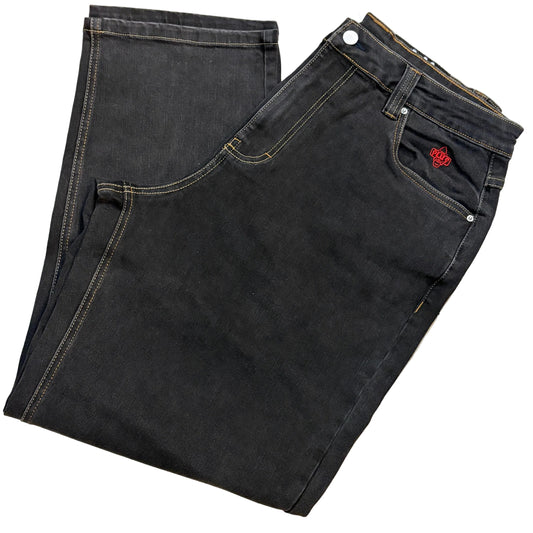 A pair of folded black denim jeans laid flat on a light wood floor, showing the front pocket, button waist, and a small red embroidered logo on the pocket.