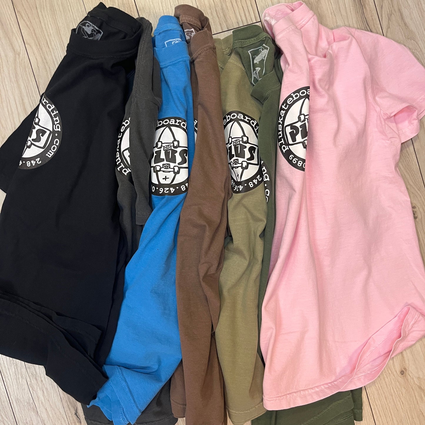 Stack of folded clothes including a pink shirt, green pants, brown jacket, blue shirt, and black pants on a wooden floor.